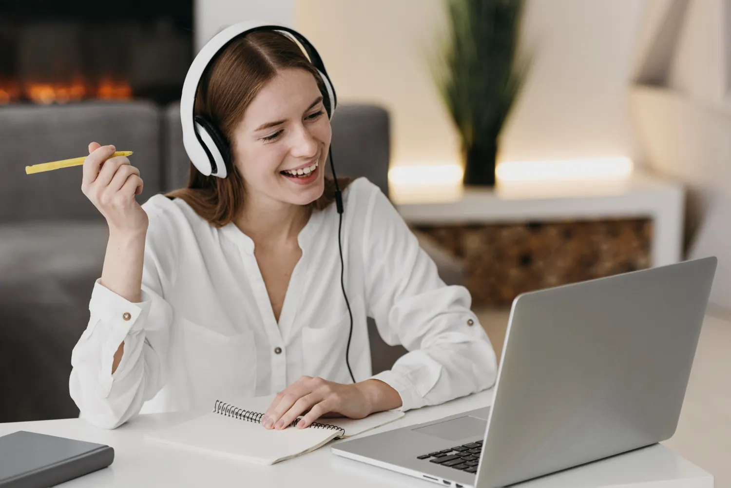 Mujer joven con auriculares sonriente mientras toma notas en un cuaderno frente a una laptop en un ambiente moderno y acogedor.