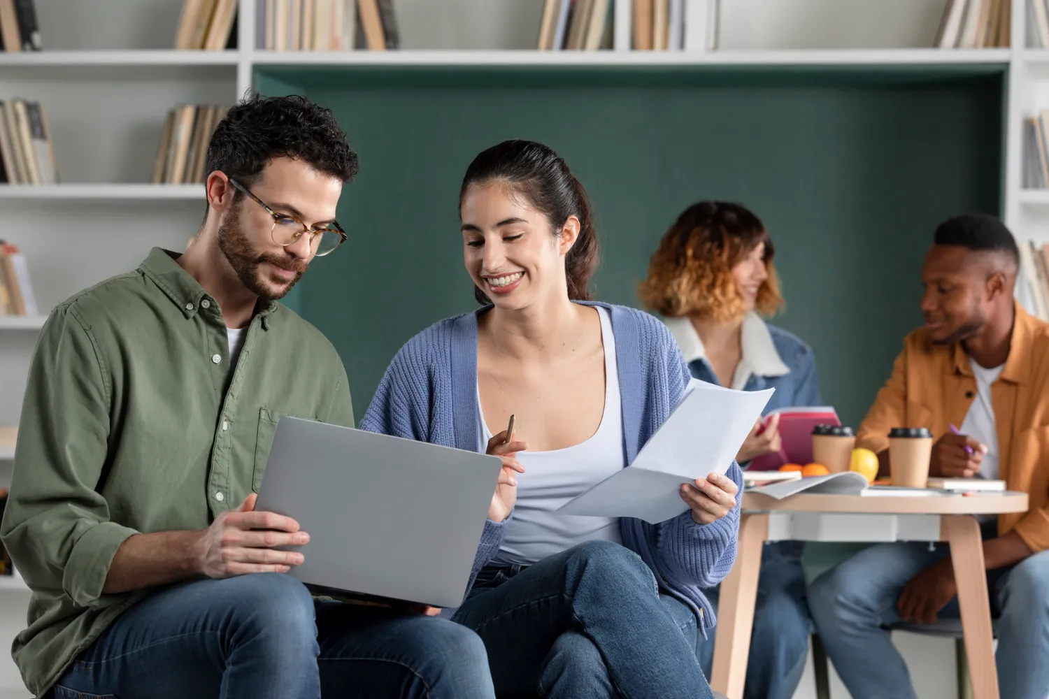 Grupo de personas colaborando en un ambiente de trabajo, revisando documentos y utilizando una computadora portátil, con estanterías de libros al fondo.