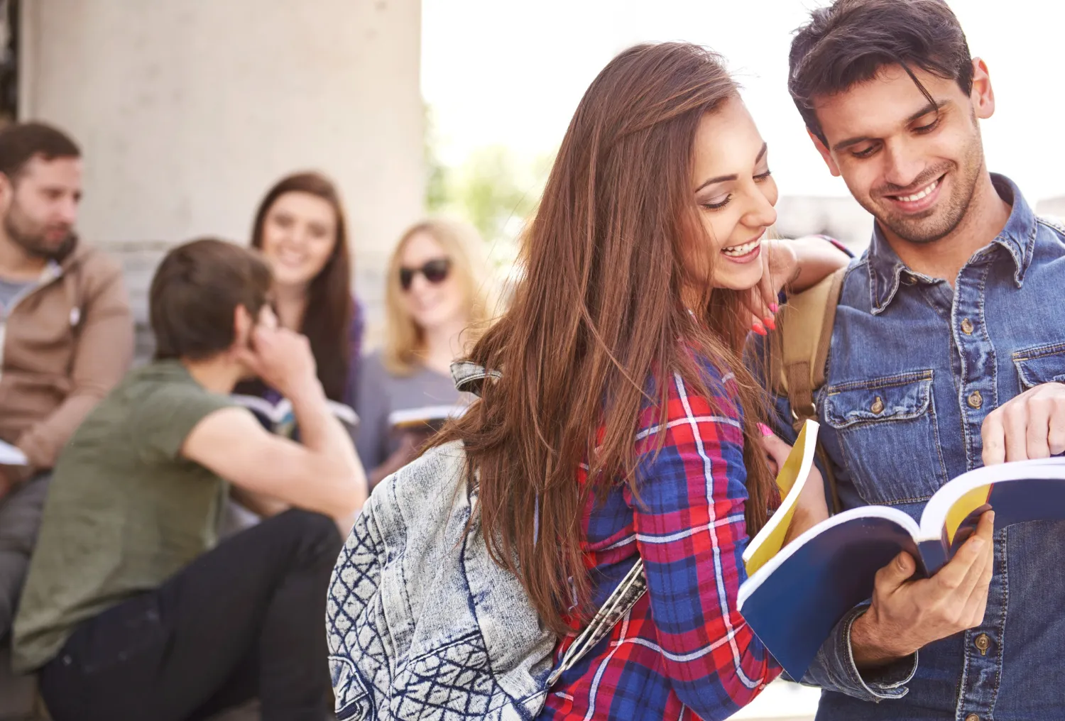 Grupo de estudiantes sonrientes en un ambiente al aire libre, revisando libros y compartiendo ideas. En primer plano, una joven con una camisa de cuadros y una mochila interactúa con un joven que sostiene un libro abierto. Al fondo, otros estudiantes conversan y disfrutan de su tiempo juntos.